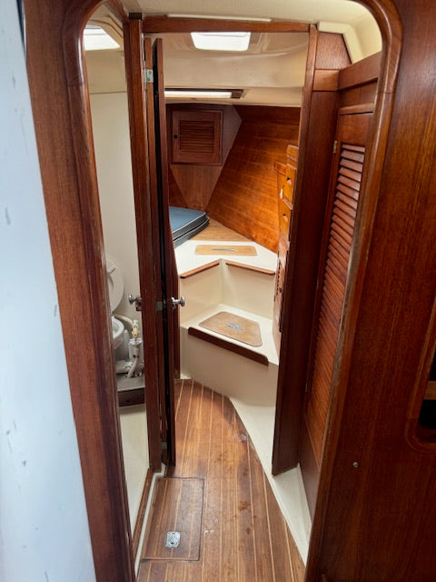 A view through a wooden doorway into a compact boat cabin interior with wood flooring, wood-paneled walls, a small bench or seating area, and a toilet visible on the left.