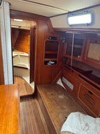 The interior of a wooden sailboat cabin with wood-paneled walls, a wooden berth at the side, a wooden table in the foreground, and a white canvas cover on the berth floor.