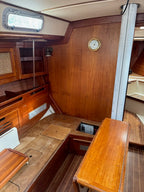 The interior cabin of a wooden boat featuring warm brown wood paneling, a wooden table, a wooden bench, and a round wall clock.
