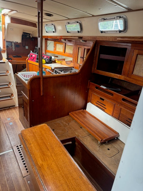 The interior cabin of a wooden sailboat or yacht, featuring warm brown wood cabinetry, a galley kitchen area, a bench seat, and small oval windows along the ceiling.