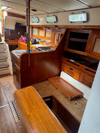 The interior cabin of a wooden sailboat or yacht, featuring warm brown wood cabinetry, a galley kitchen area, a bench seat, and small oval windows along the ceiling.