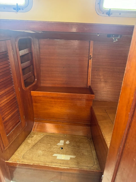 An interior wooden cabin space with wood-paneled walls, a bench or seat, a small shelf, and a floor hatch, photographed from inside the compartment.