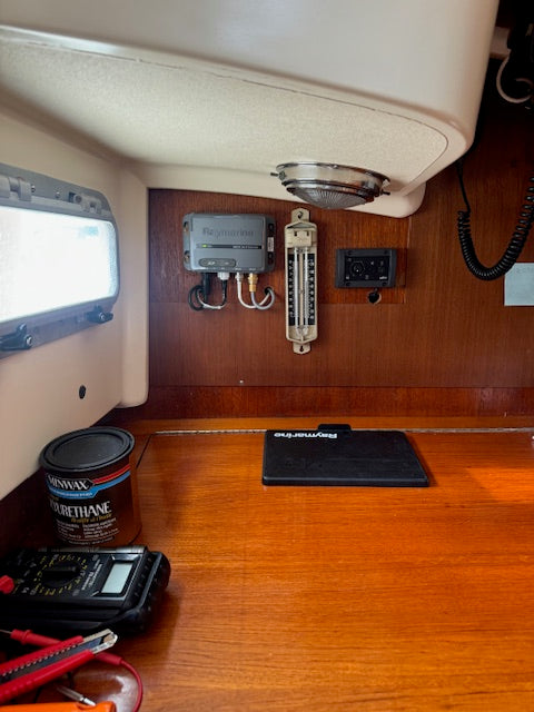 A close-up view of a wooden boat cabin interior showing a wood-grain workbench, an electronic control panel with a thermometer and gauges, a ceiling light, and various tools on the table.