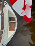 A close-up, high-angle view of a boat's corner with a cream-colored hull, wooden deck flooring, metal railings, and a Canadian flag hanging over the side, with dark water below.