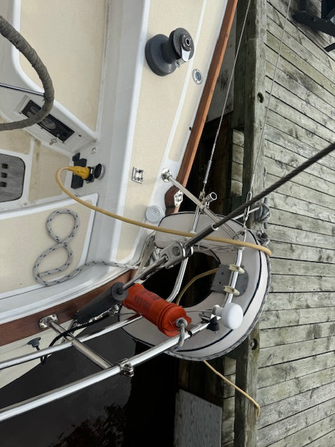 A close-up, angled view of a boat's deck and rigging next to a wooden dock, featuring a white and beige hull, winch system, ropes, and an orange handle.