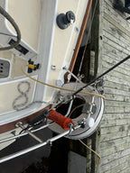 A close-up, angled view of a boat's deck and rigging next to a wooden dock, featuring a white and beige hull, winch system, ropes, and an orange handle.