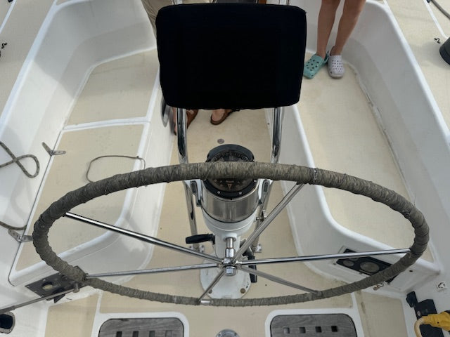 A top-down view of a boat helm station featuring a large metal steering wheel with a gray wrapped rim, a black captain's chair, and a person's legs and feet visible on the cream-colored deck below.