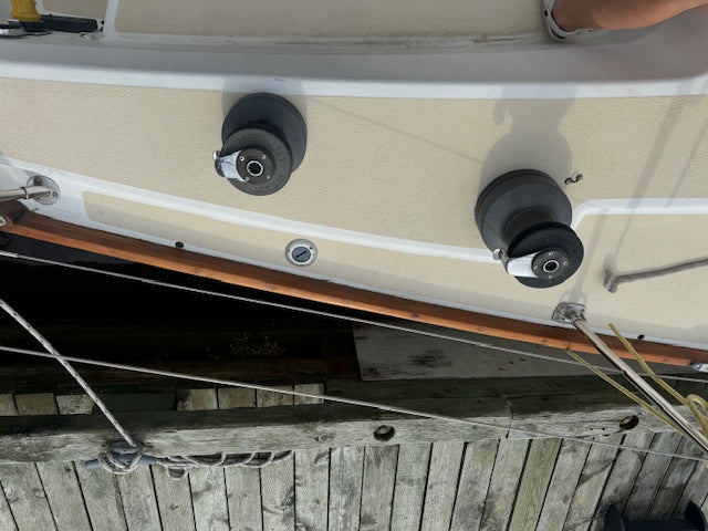 Close-up of a boat deck with two black winches mounted on a beige fiberglass surface, alongside a wooden railing and ropes at the dock.