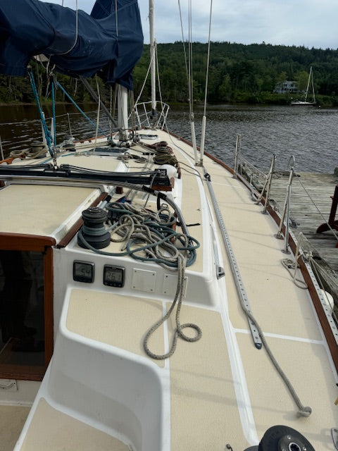 The deck of a sailboat docked at a marina, showing a beige fiberglass deck, coiled ropes, a blue canvas cover, and a background of water and forest.