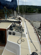 The deck of a sailboat docked at a marina, showing a beige fiberglass deck, coiled ropes, a blue canvas cover, and a background of water and forest.