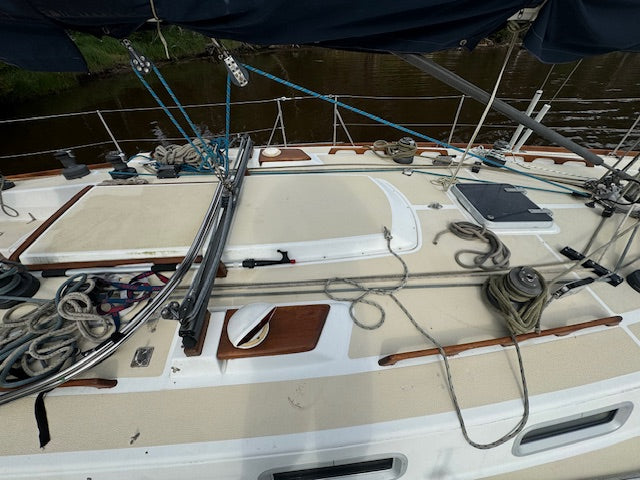 The deck of a sailboat on water, showing the white and beige fiberglass deck, ropes, rigging, winch, and a dark blue canvas cover overhead.