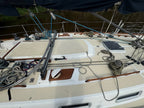 The deck of a sailboat on water, showing the white and beige fiberglass deck, ropes, rigging, winch, and a dark blue canvas cover overhead.