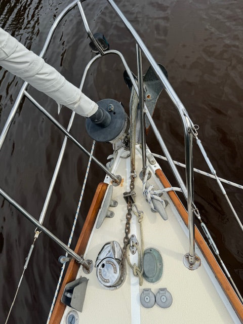 A top-down view of a sailboat's deck near the bow, showing rigging, cleats, a chain, and a white sailcloth extending from the mast, with dark water surrounding the boat.