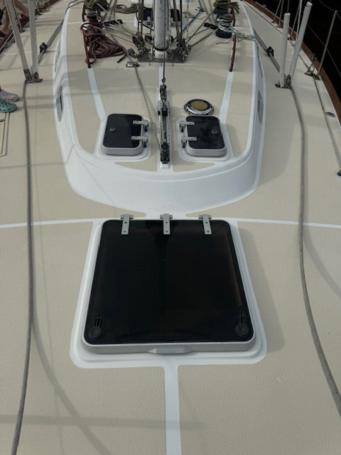 A close-up view of a black rectangular hatch cover with metal latches set into a white recessed panel on a beige deck, with boat rigging and equipment visible above.