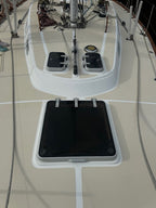 A close-up view of a black rectangular hatch cover with metal latches set into a white recessed panel on a beige deck, with boat rigging and equipment visible above.