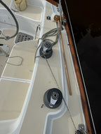 Close-up view of a boat deck showing two black and silver winches with coiled rope, beige deck flooring, a wooden cleat rail, and dark water at the side.