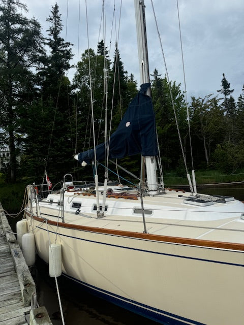 A cream-colored sailboat docked at a wooden pier with trees in the background under an overcast sky. The boat has a dark blue cover on its mast and white fenders attached to the side.