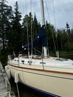A cream-colored sailboat docked at a wooden pier with trees in the background under an overcast sky. The boat has a dark blue cover on its mast and white fenders attached to the side.
