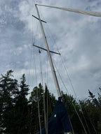 A close-up low-angle view of a tall wooden mast and rigging on a sailboat, with a dark blue canvas cover at the base, set against a cloudy sky and trees.