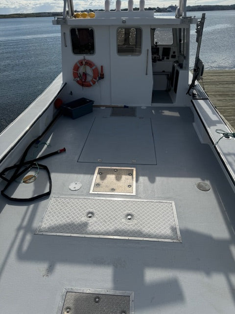 The deck of a white fishing boat docked at a pier, showing the gray floor, metal hatch covers, a black coiled hose, and an orange life preserver near the cabin.