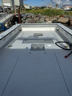 The rear deck of a white boat, showing a gray composite floor with hatch covers, a black coiled rope, and an outdoor dockside background with rocks and grass.
