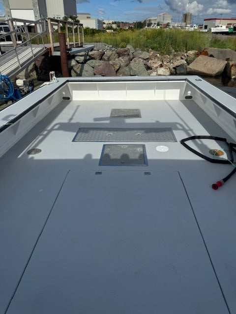 The rear deck of a white boat, showing a gray composite floor with hatch covers, a black coiled rope, and an outdoor dockside background with rocks and grass.