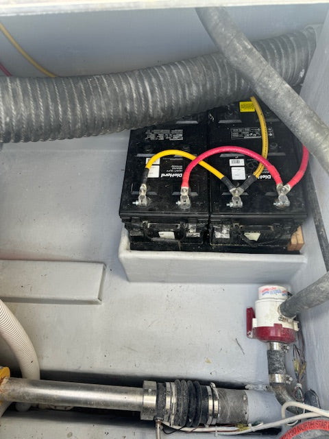 Two black marine batteries with red and yellow cables connected in a gray compartment with hoses and pipes.