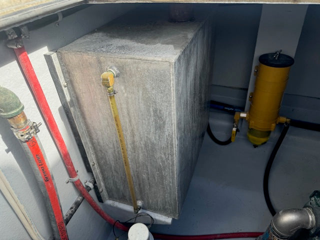 A large rectangular gray concrete or metal utility tank with a yellow vertical pipe attached, photographed inside a confined mechanical space alongside red and black hoses and a yellow cylindrical filter unit.