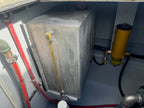 A large rectangular gray concrete or metal utility tank with a yellow vertical pipe attached, photographed inside a confined mechanical space alongside red and black hoses and a yellow cylindrical filter unit.
