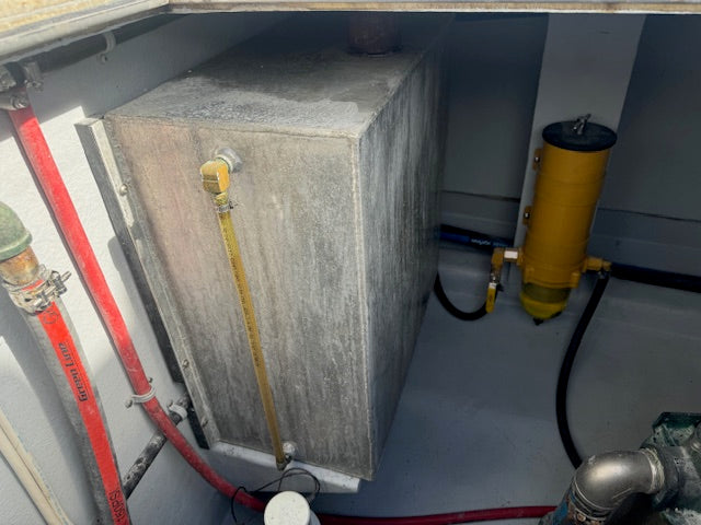 A large rectangular gray concrete or metal utility box with a yellow vertical pipe attached, photographed inside a confined mechanical compartment alongside red and black hoses and a yellow cylindrical component.