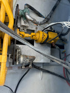 A close-up view of industrial machinery showing a yellow hydraulic or mechanical component connected by hoses and cables to other machinery parts inside a gray metal housing.