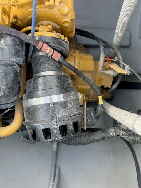 A used black alternator with a silver metal band, mounted on a yellow industrial engine with various hoses and wires visible.