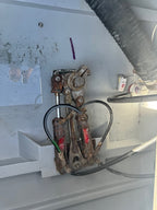 A close-up of a used mechanical hydraulic or steering linkage assembly with rusty metal parts, black hoses, and a small purple marker on a light gray painted surface.