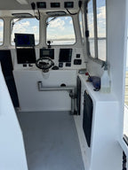 The helm station of a boat cabin with white walls, a steering wheel, multiple electronic navigation screens, and a view of water and sky through the windows.