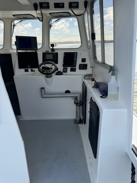 The helm station of a boat cabin with white walls, a steering wheel, multiple electronic navigation screens, and a view of water and sky through the windows.