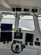 A white boat cabin interior with multiple black electronic navigation screens, a steering wheel, and windows looking out onto open water under a cloudy sky.