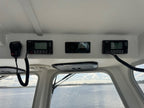 Four black marine electronic navigation and communication devices mounted on the white ceiling of a boat cabin, with windows showing water and distant land below.
