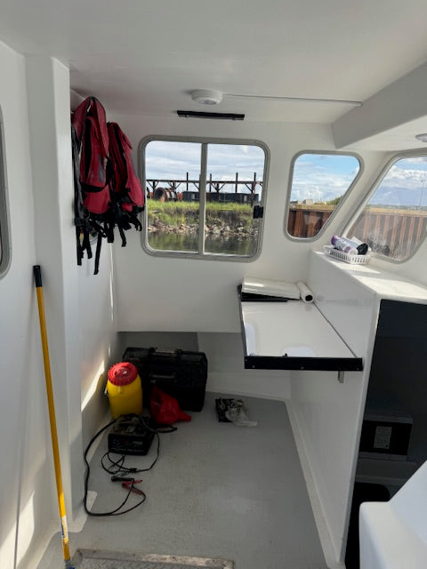 The interior cabin of a boat with white walls and windows, showing a red and black life jacket hanging on the left, a yellow pole leaning against the wall, a black storage case on the floor, and a fold-out table mounted on the side.
