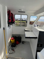 The interior cabin of a boat with white walls and windows, showing a red and black life jacket hanging on the left, a yellow pole leaning against the wall, a black storage case on the floor, and a fold-out table mounted on the side.