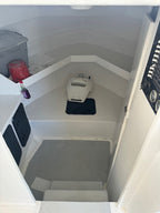 An interior view of a boat cabin showing a narrow staircase leading downward, with white walls and floor, a gray carpeted step, a small white fixture on the floor, and some storage items on a shelf.