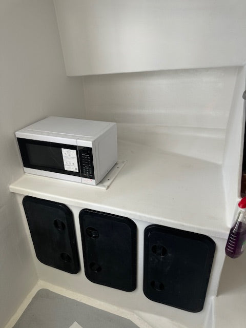 A white microwave oven sits on a white countertop in a compact kitchen or storage nook with three black storage compartments below.