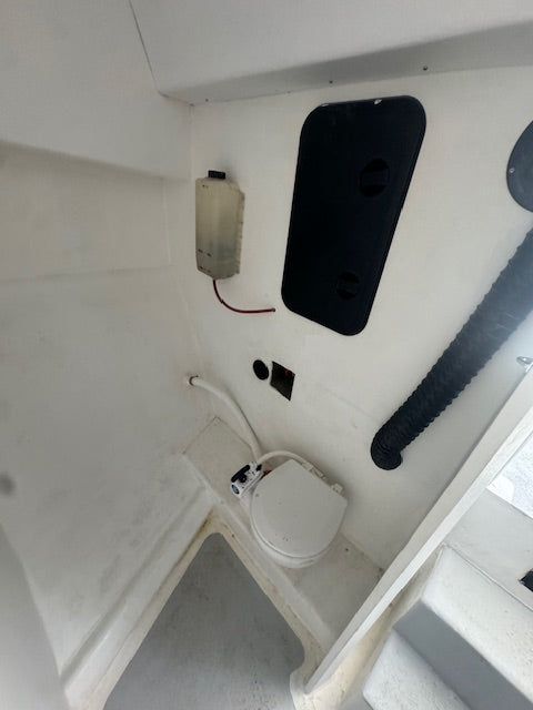 A close-up view of a white toilet fixture installed in a white boat cabin or utility space, with a black hatch cover, a black flexible hose, and a small electrical box on the wall.