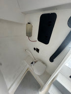 A close-up view of a white toilet fixture installed in a white boat cabin or utility space, with a black hatch cover, a black flexible hose, and a small electrical box on the wall.