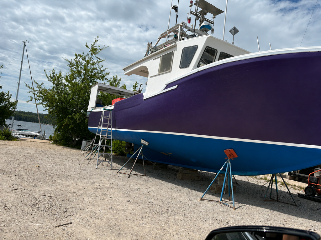A large boat in a purple and blue hull, supported by metal stands on a gravel dock with trees, water, and a cloudy sky in the background.