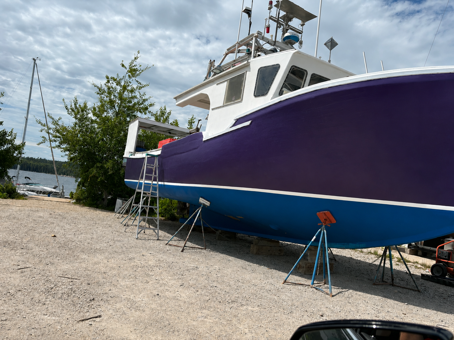 A large boat in a purple and blue hull, supported by metal stands on a gravel dock with trees, water, and a cloudy sky in the background.