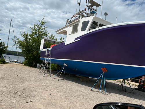 A large boat in a purple and blue hull, supported by metal stands on a gravel dock with trees, water, and a cloudy sky in the background.
