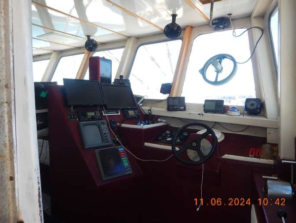 The interior of a boat's navigation deck, featuring a dark red control console with multiple electronic screens, a black steering wheel, and a white bridge cabin with large windows. A digital timestamp is visible in the bottom right corner.