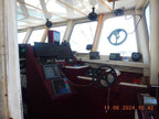 The interior of a boat's navigation deck, featuring a dark red control console with multiple electronic screens, a black steering wheel, and a white bridge cabin with large windows. A digital timestamp is visible in the bottom right corner.