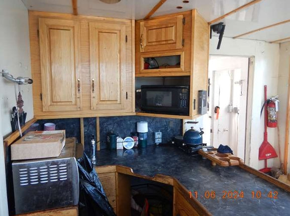 A compact kitchen corner with light wood cabinets, a black microwave, a dark countertop, and various household items including a blue kettle, a cardboard box, and cleaning supplies.