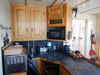 A compact kitchen corner with light wood cabinets, a black microwave, a dark countertop, and various household items including a blue kettle, a cardboard box, and cleaning supplies.